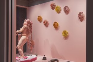 In a street-level window gallery space, which has a pale pink painted floor and walls, a life-size pink ceramic female figure stands on a low circular plinth that is covered in white and red fake fur. On the floor around the figure are several dark green and blue snakes. The body and head of the figure is covered in clay forms of flowers and leaves, the figure has long pink hair that almost reaches the floor. The hands are holding pink hair strands which have snake heads. On the wall behind the figure are several yellow ochre, orange and pink ceramic flower heads.]