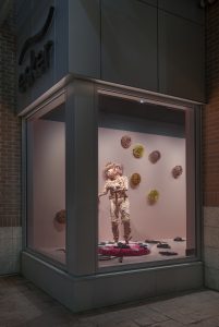 In a street-level window gallery space, which has a pale pink painted floor and walls, a life-size pink ceramic female figure stands on a low circular plinth that is covered in white and red fake fur. On the floor around the figure are several dark green and blue snakes. The body and head of the figure is covered in clay forms of flowers and leaves, the figure has long pink hair that almost reaches the floor. The hands are holding pink hair strands which have snake heads. On the wall behind the figure are several yellow ochre, orange and pink ceramic flower heads.]