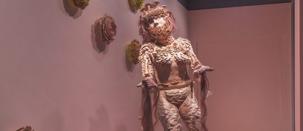 In a street-level window gallery space, which has a pale pink painted floor and walls, a life-size pink ceramic female figure stands on a low circular plinth that is covered in white and red fake fur. On the floor around the figure are several dark green and blue snakes. The body and head of the figure is covered in clay forms of flowers and leaves, the figure has long pink hair that almost reaches the floor. The hands are holding pink hair strands which have snake heads. On the wall behind the figure are several yellow ochre, orange and pink ceramic flower heads.]