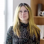 A head and shoulders view of the artist Yvonne Kustec, her hair is long and is yellow and blond, she has round glasses and is looking to the upper right of the camera. She is wearing a black long sleeved polo neck top with with a pattern of multiple small blue, dark red, pale yellow, green, white and pink flowers. In the background, part of a bookshelf is visible and also a white window frame.